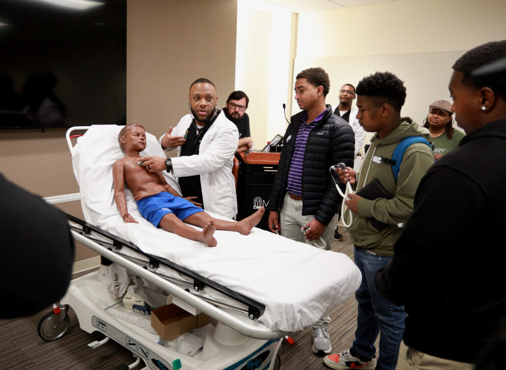 A man lying on a hospital bed, surrounded by medical personnel and onlookers. The room is filled with people, some standing around the bed, others observing from a distance. The atmosphere seems tense.