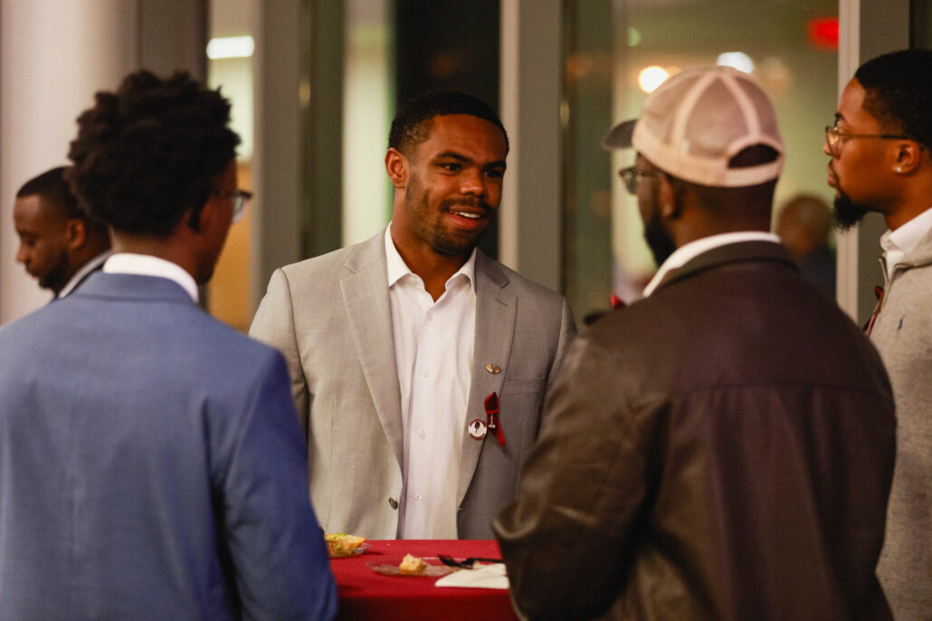 A group of men in suits gathered around a table. One man is standing and smiling while others stand or sit behind him. They are indoors, possibly at an event.