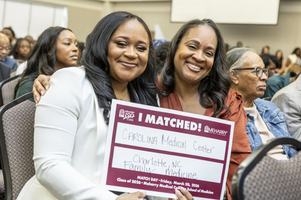 Two women posing with a sign at an event. One woman is holding a cardboard sign that says "I MATCHED!" and the other is smiling. They are standing in front of a crowd. The setting appears to be indoors, possibly a conference or ceremony.