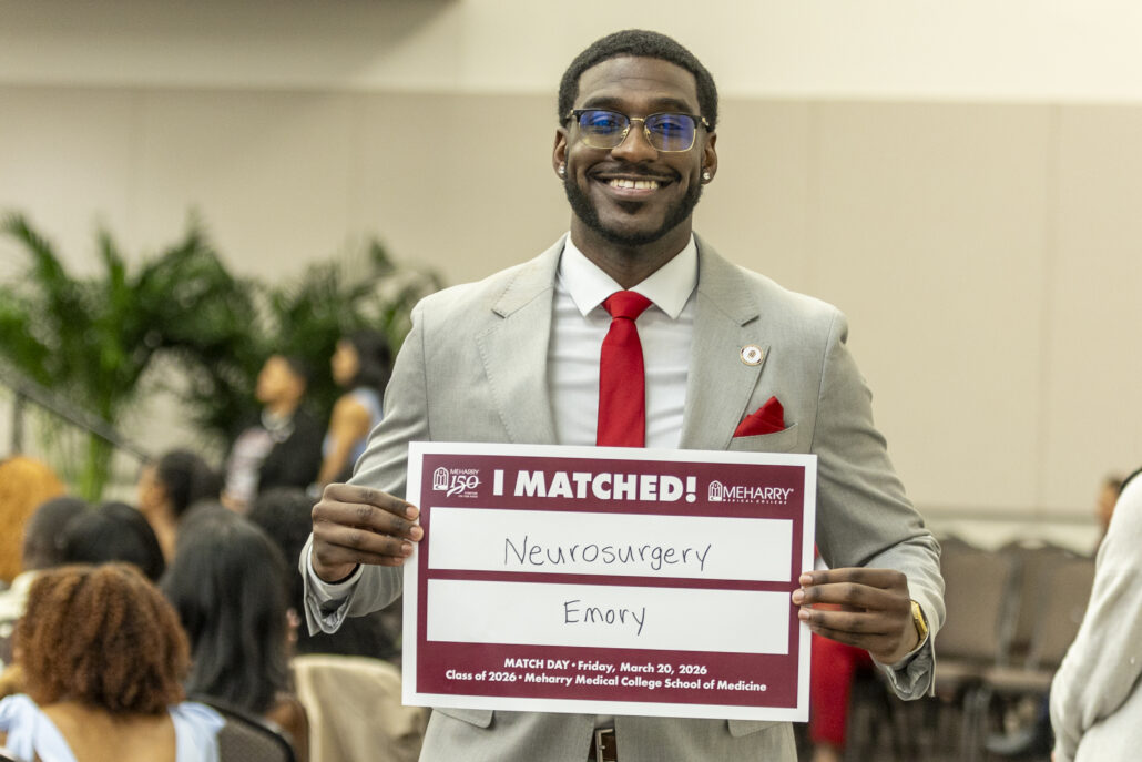 Man holding sign at event. Sign says "I MATCHED NEWBORN SURGERY". Man wearing red tie and glasses.