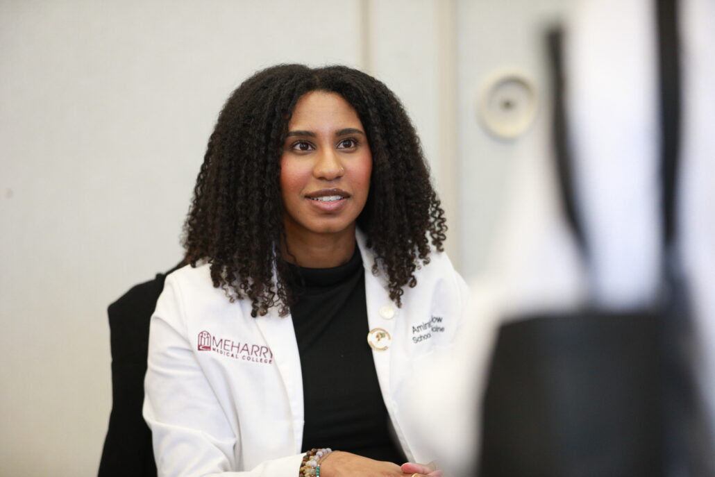 Woman sitting at desk, smiling, looking to her right. She has dark hair, wears glasses, a black shirt, and a white coat. In front of her is a blurred image of another person's face. The setting appears to be an office or classroom.